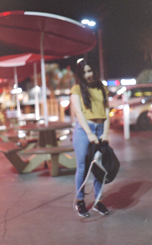 Film Photo of Gabriela Inzunza a 21-year-old in CA posing outside the local In-n-out at night. She wears a yellow-colored t-shirt and blue jeans.